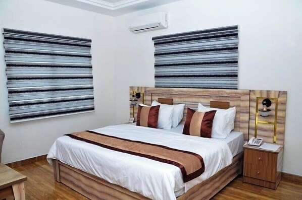 Un dormitorio con una cama grande, un cabecero de madera, una mesita de noche con lámpara y dos ventanas amplias con persianas.