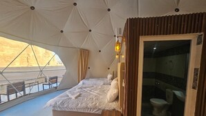 A tented room with a bed, a small wooden table, and a bathroom visible through a glass door. - Wadi Rum Desert Camp (Wadi Rum)