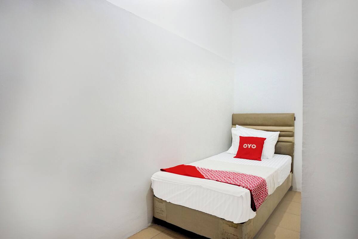 Un letto piccolo e ben rifatto con copriletto bianco e rosso, una testiera beige e un solo cuscino rosso con la scritta "OYO".