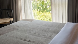 A bed with a white sheet in a room with a large window, curtains, and a chair. - TERRATONE BOUTIQUE BUSINESS HOTEL (Kollam)