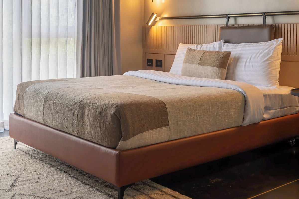 Une chambre d’hôtel moderne avec un grand lit, une table de chevet, une lampe et une vue sur l’extérieur à travers des rideaux transparents.