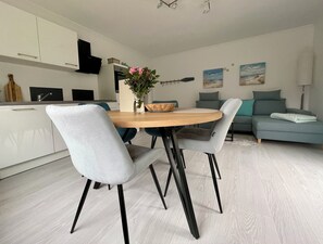 Une cuisine moderne avec des meubles blancs, une table à manger avec des chaises et un coin salon avec un canapé et des coussins.