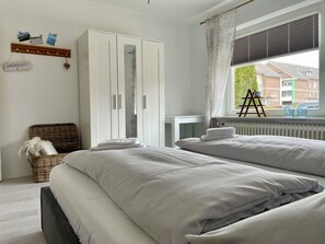 Une chambre avec deux lits, une armoire, une chaise en osier, un radiateur et une fenêtre donnant sur des immeubles.