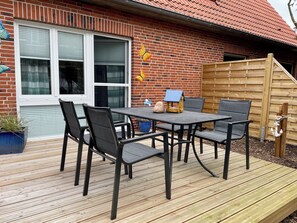 Une terrasse avec une table et des chaises en bois, une petite maquette d’une maison bleue et un mur de briques avec une fenêtre.