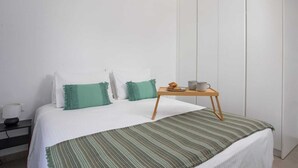 Un dormitorio con una cama, almohadas, una mesita de noche con una taza y un plato, y una lámpara.