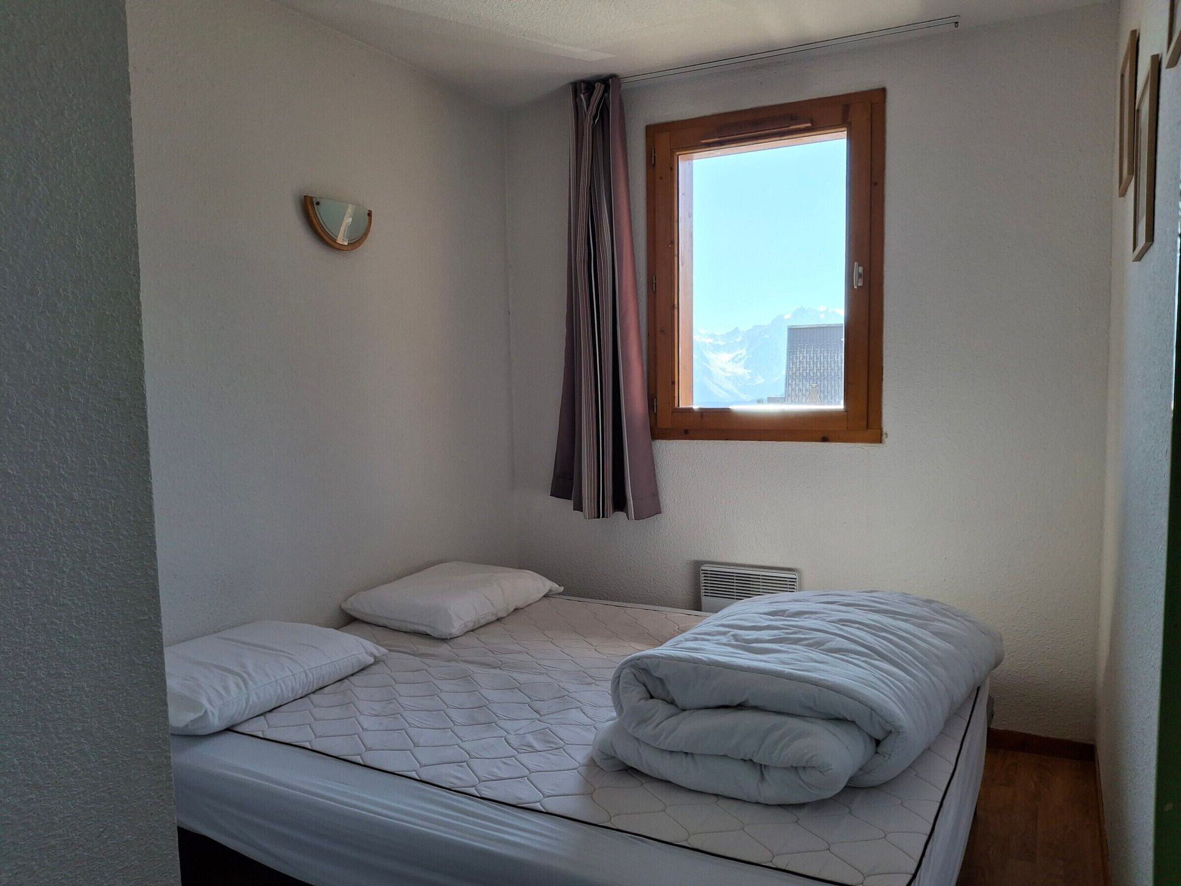 A small, simple bedroom with a single bed, a window with curtains, and a wall-mounted heater.