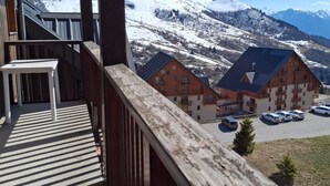 A wooden balcony with a table, overlooking a snow-covered mountain and a row of buildings with solar panels. - Comfortable 2-BR Apt at the Foot of the Slopes (Saint-François-Longchamp)
