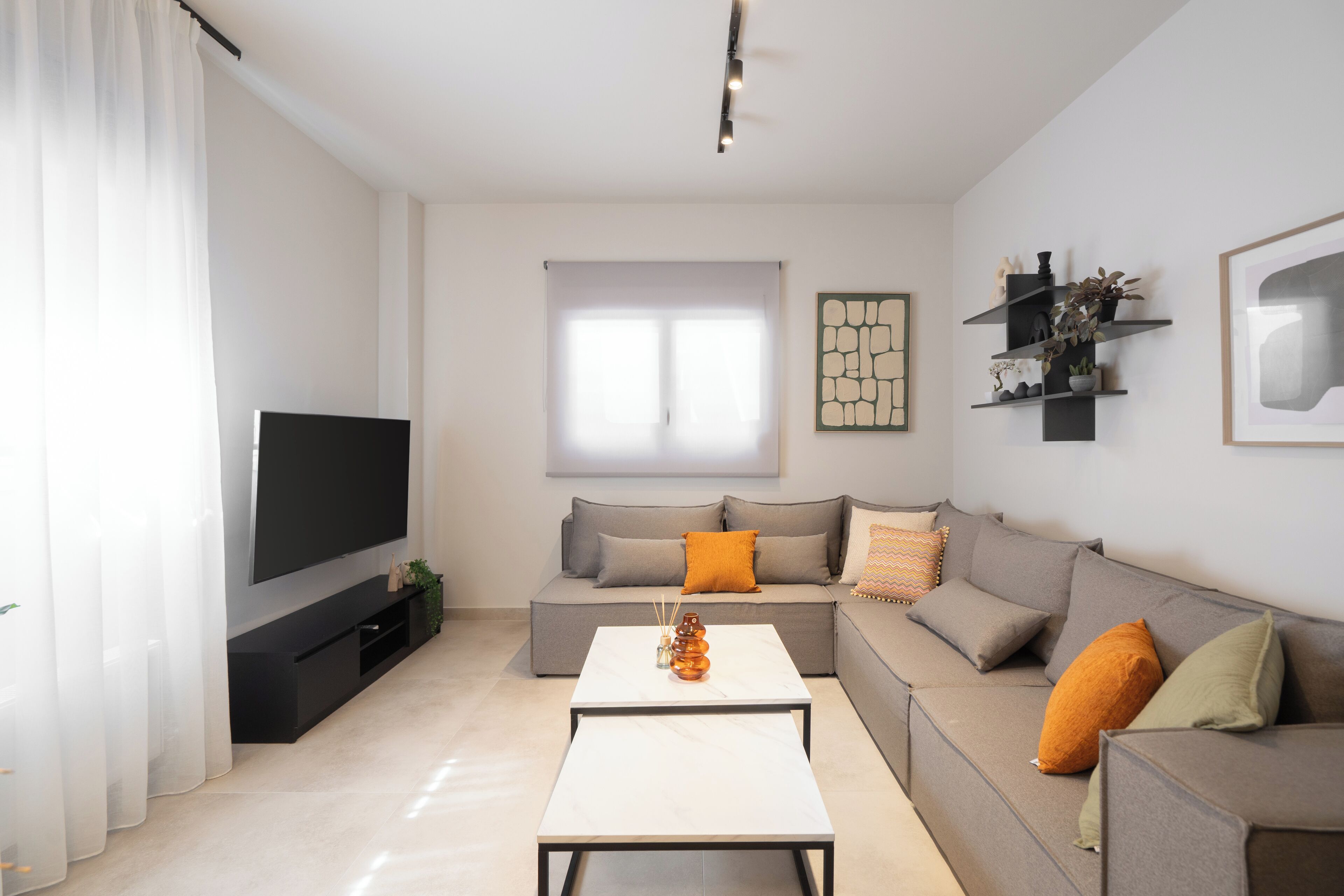A modern living room with a flat-screen TV, a sofa, a coffee table, and a window with sheer curtains.