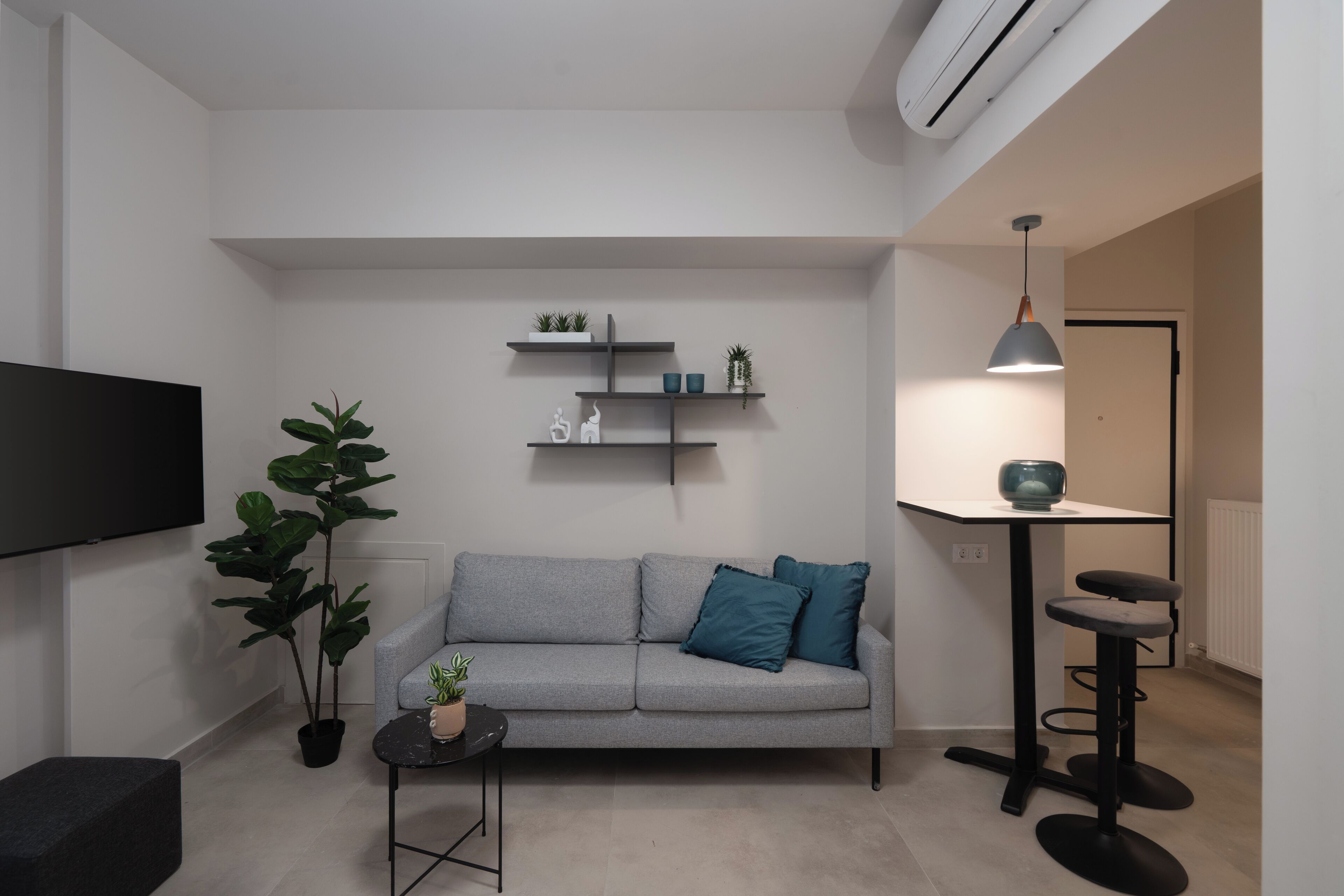 Ein modernes Wohnzimmer mit einem grauen Sofa, einem kleinen runden Tisch, einer Topfpflanze und einem an der Wand montierten Fernseher.