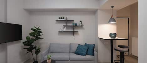 Ein modernes Wohnzimmer mit einem grauen Sofa, einem kleinen runden Tisch, einer Topfpflanze und einem an der Wand montierten Fernseher.