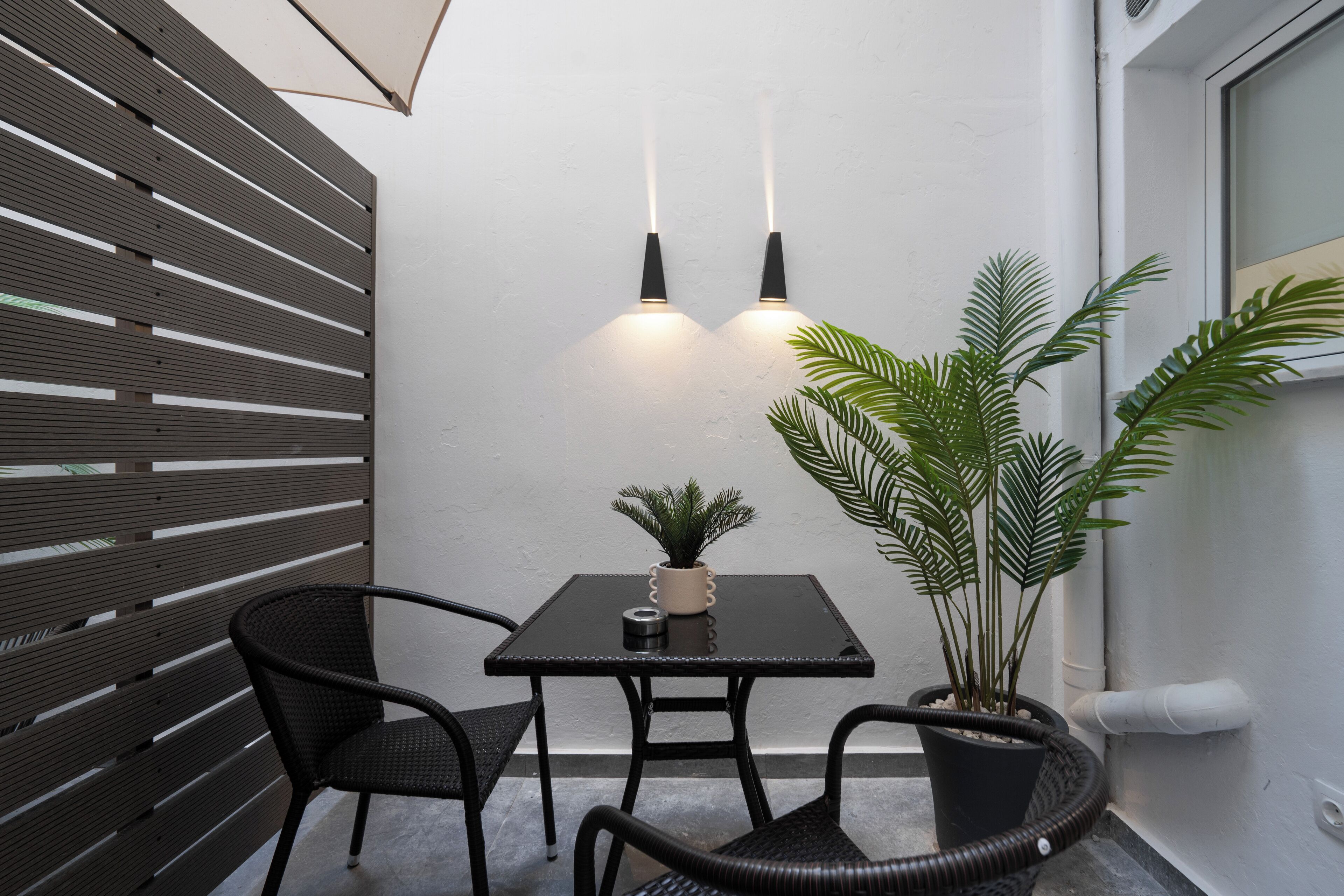 A small outdoor seating area with a table and two chairs, a potted plant, and two wall-mounted lights.