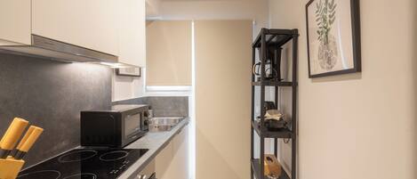 A compact kitchen with a black induction cooktop, a microwave, and a stainless steel sink. There is a wooden knife block on the counter, and a framed plant on the wall.