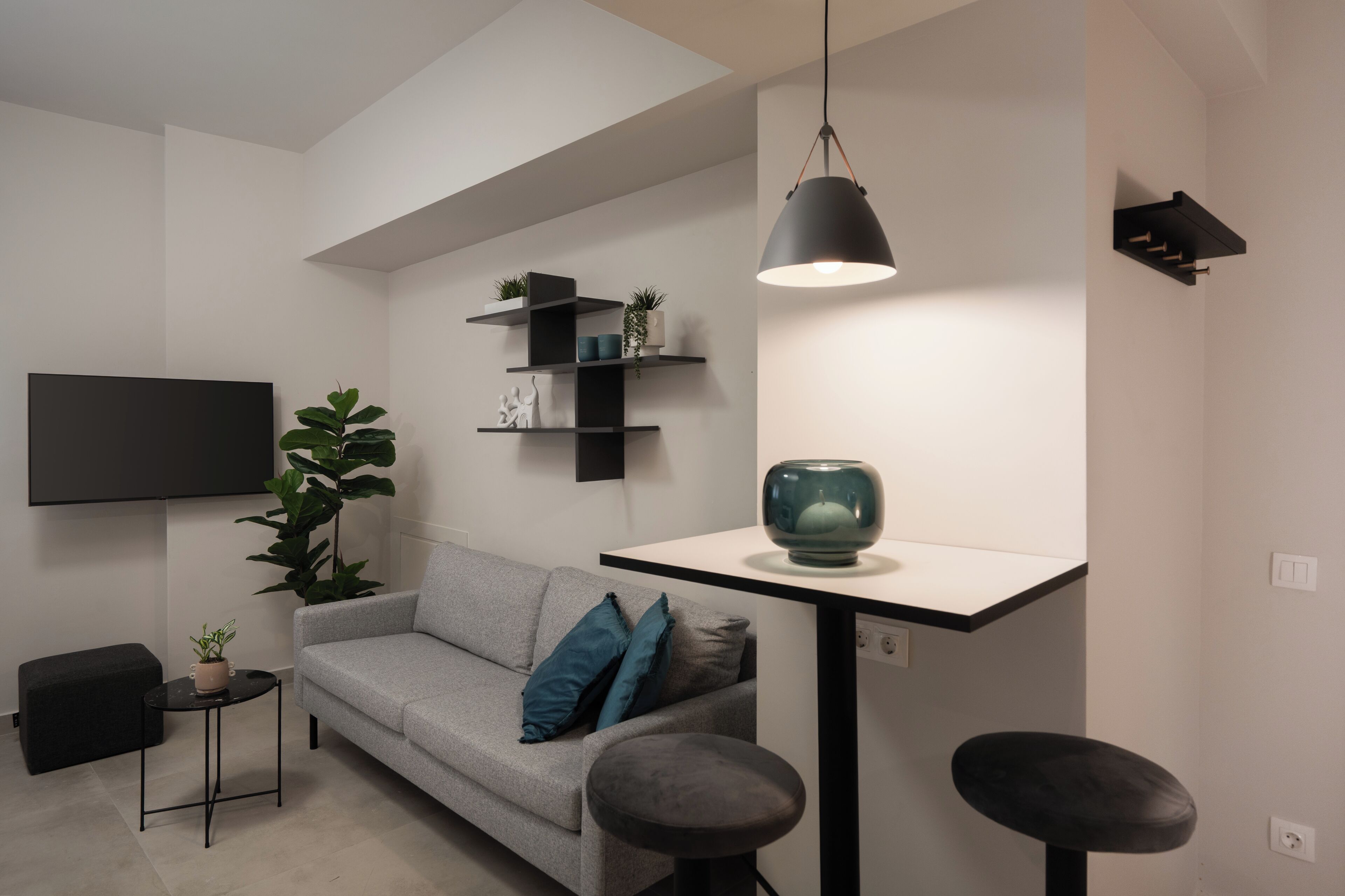 Una sala de estar moderna con un sofá gris, una mesita redonda pequeña, una lámpara colgante negra y un televisor montado en la pared.