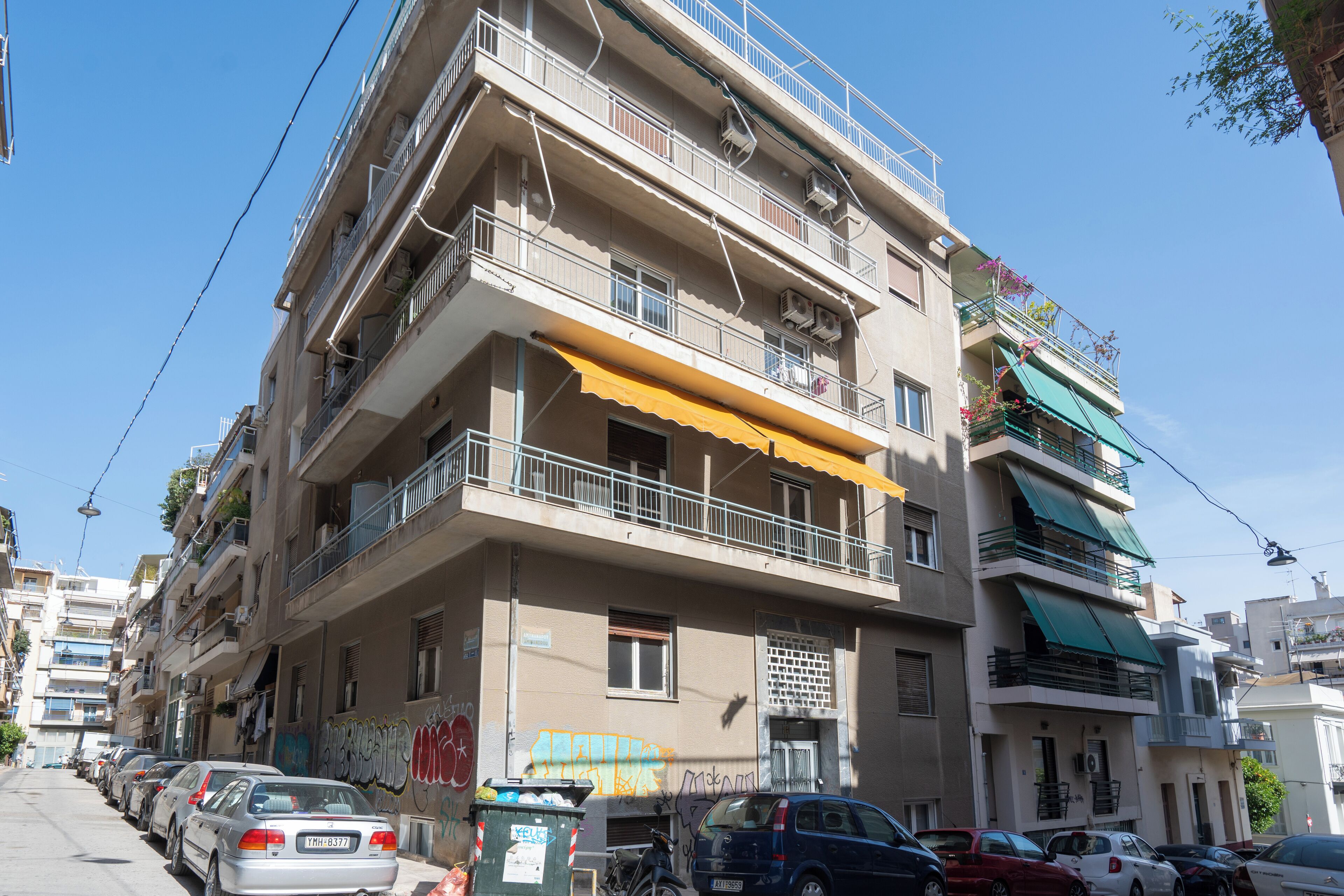 A multi-story residential building with balconies, a row of parked cars, and a visible street sign.
