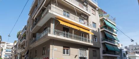 A multi-story residential building with balconies, a row of parked cars, and a visible street sign.
