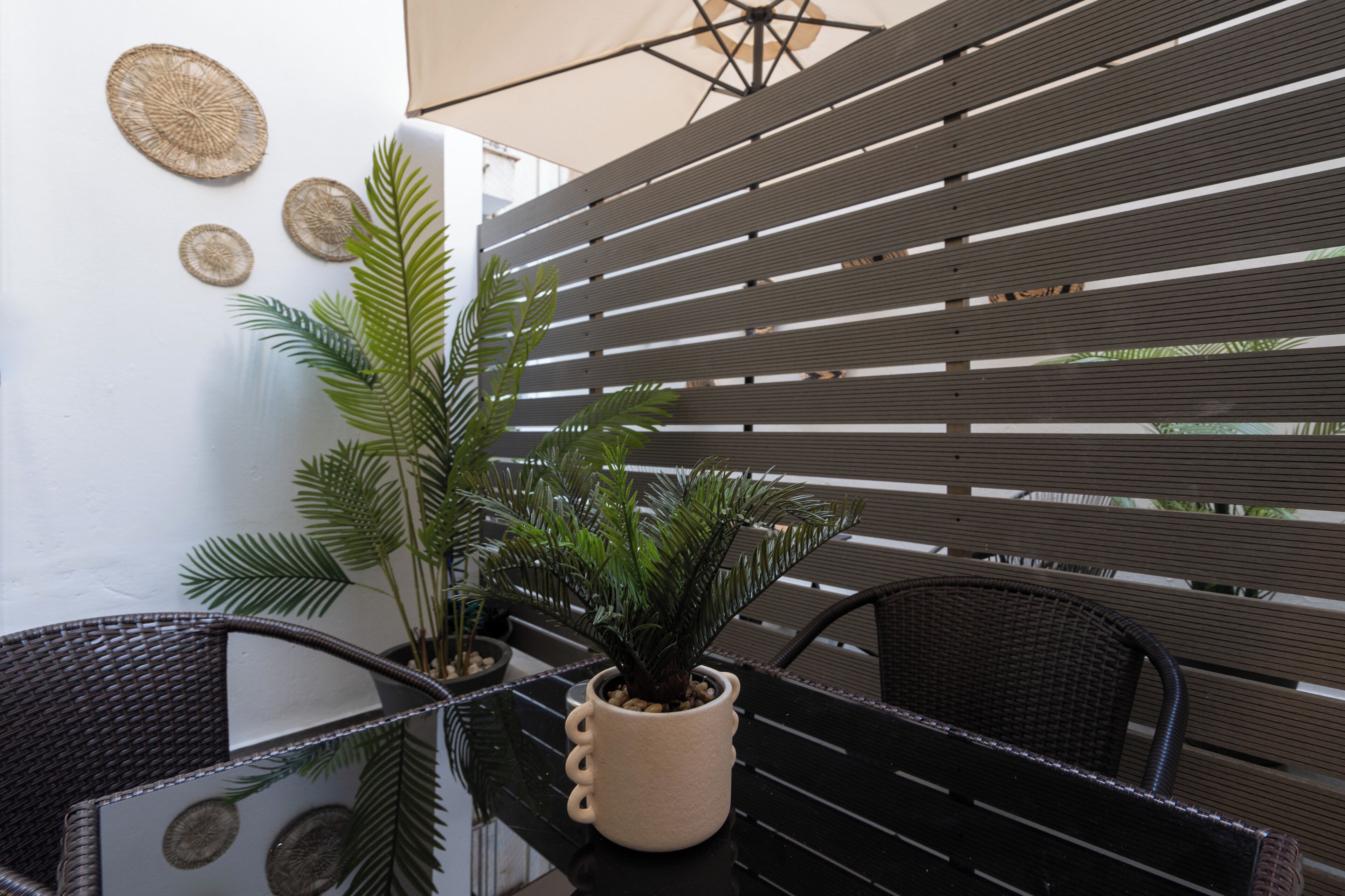 A balcony with a glass table, two wicker chairs, a potted plant, and a wooden slatted wall.
