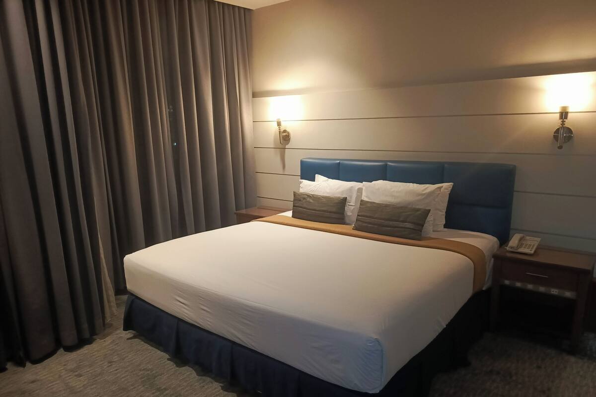 Quarto de hotel com uma cama grande, cabeceira, duas mesas de cabeceira, telefone e cortina.