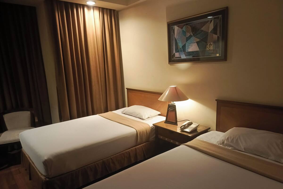 Quarto de hotel com duas camas, uma mesa de cabeceira com abajur, telefone e um quadro na parede.