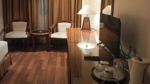 A hotel room with a wooden floor, a desk with a lamp, a chair, a bed, and a large window with curtains.