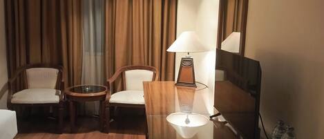 Ein Hotelzimmer mit Holzboden, einem Schreibtisch mit Lampe, einem Stuhl, einem Bett und einem großen Fenster mit Vorhängen.