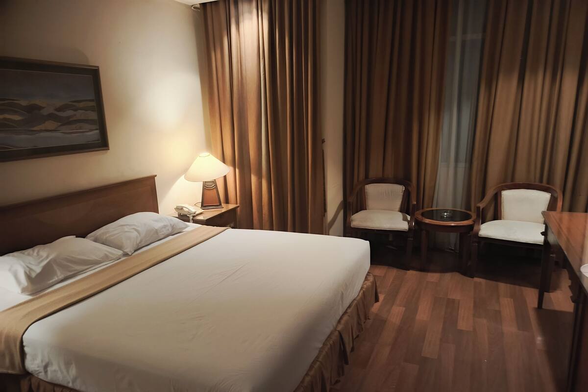Quarto de hotel com uma cama grande, uma mesa de cabeceira com abajur, duas cadeiras e uma mesinha.