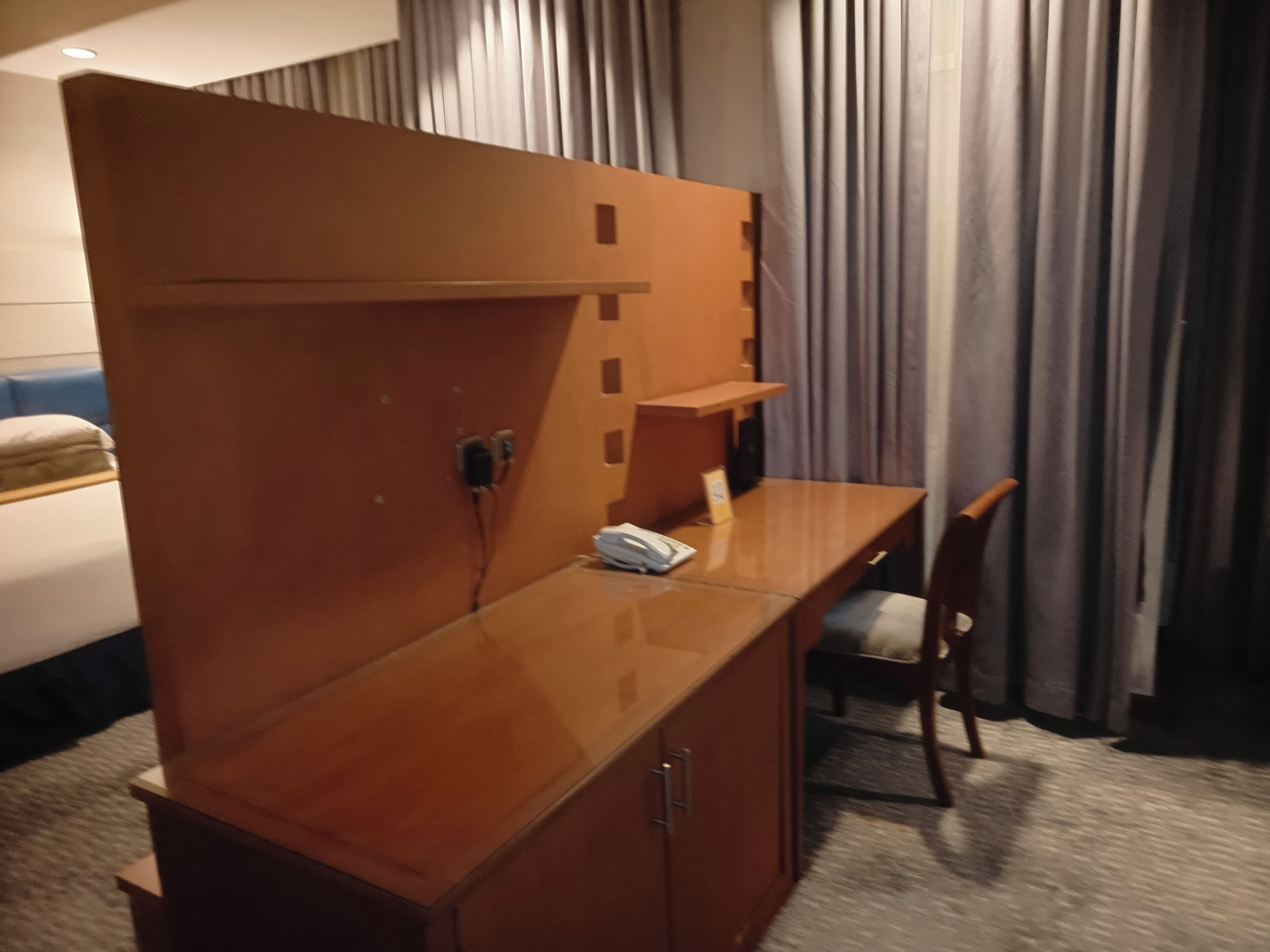 A hotel room with a wooden desk, a chair, a telephone, and a bed with a blue headboard.