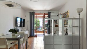 Una sala de estar moderna con una mesa de comedor de vidrio, sillas blancas, un televisor, un mueble con puertas de vidrio y un balcón con vistas.