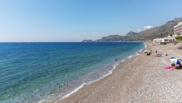 Una playa de guijarros con agua cristalina, algunas personas y edificios al fondo.