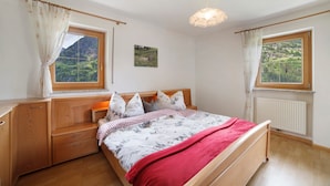 Une chambre à coucher avec un lit, des meubles en bois, deux fenêtres avec des rideaux, un radiateur et un tableau au mur.