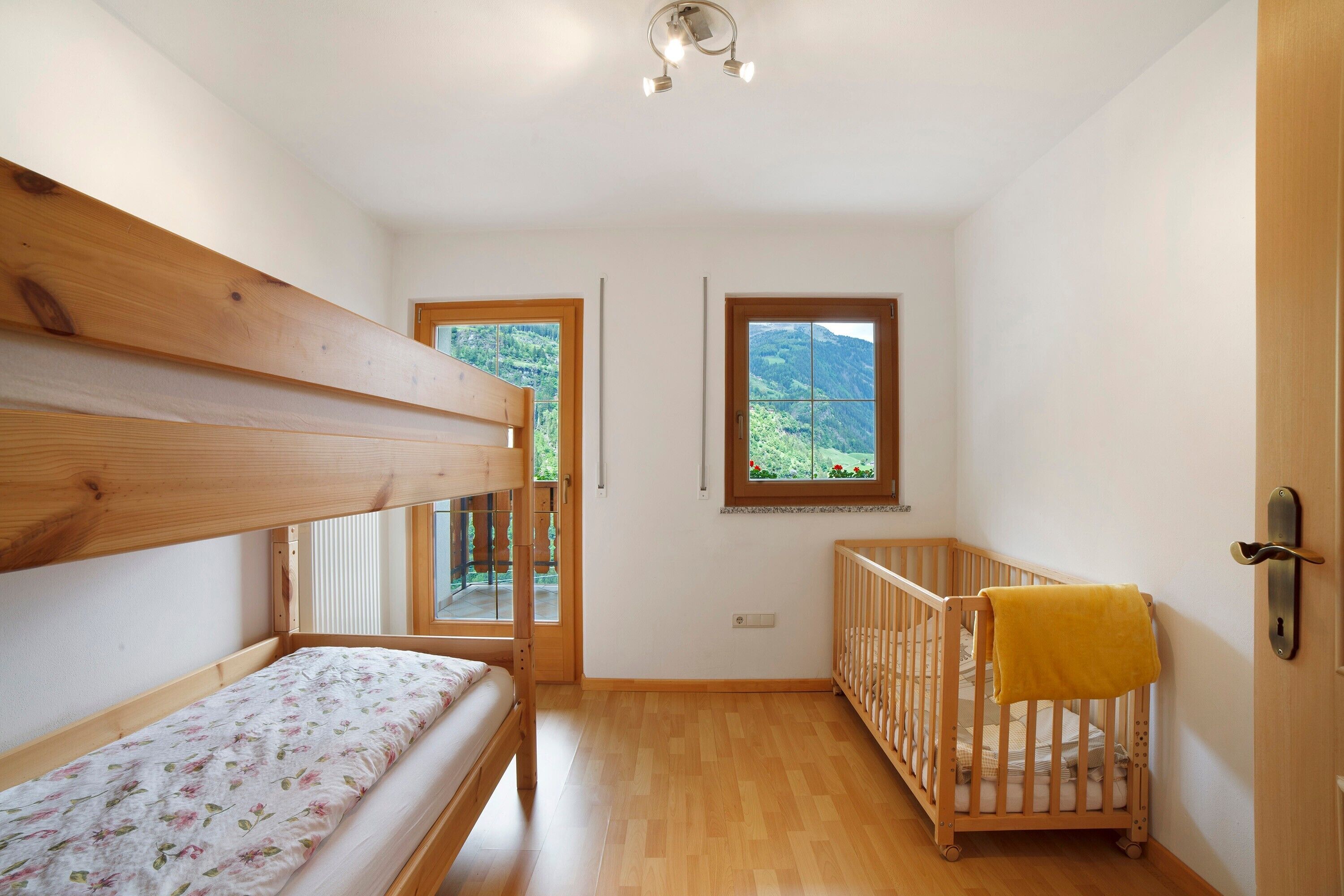 Una habitación con una cama litera, una cuna, un ventanal con vistas y una puerta con manija.
