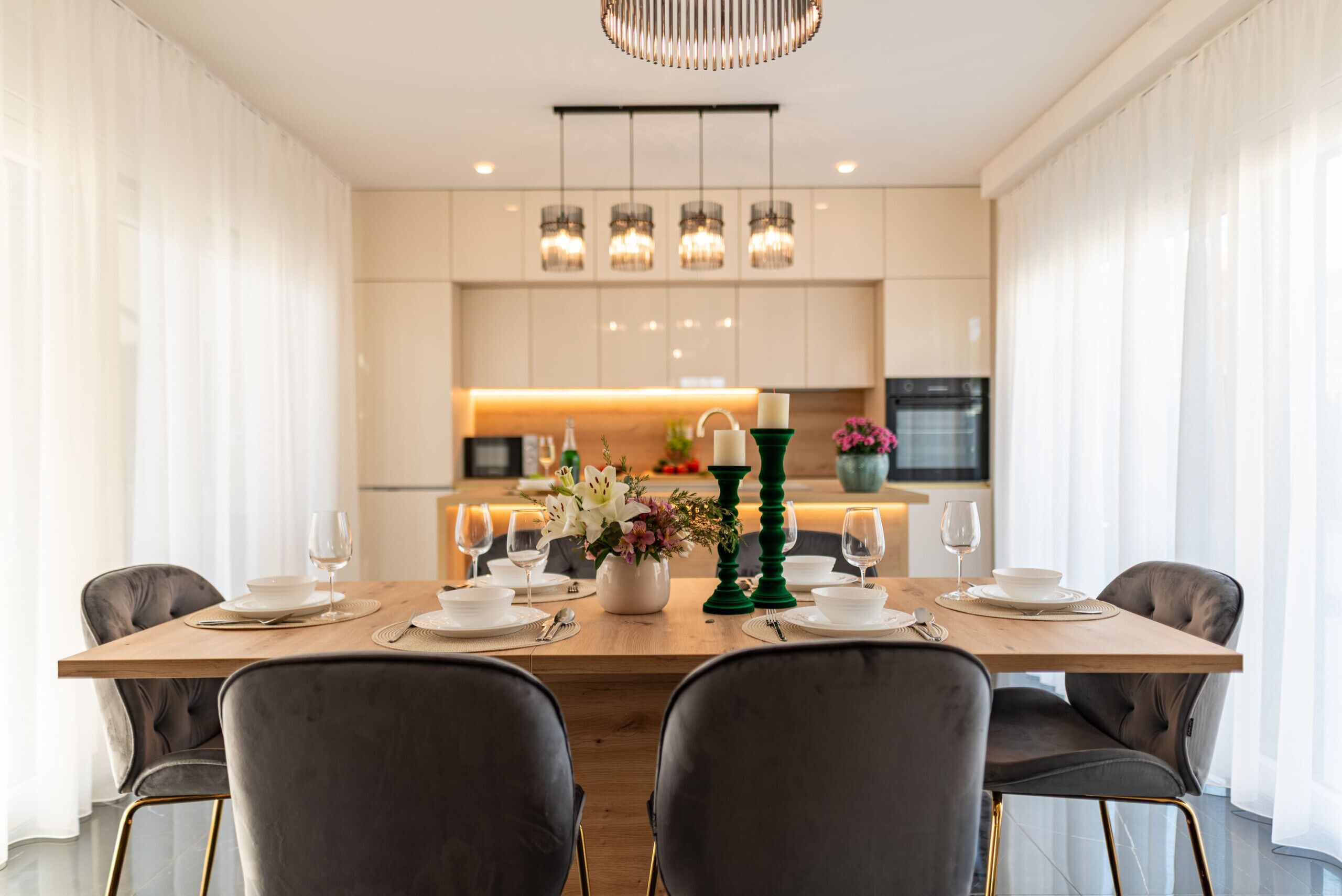Uma área de jantar moderna com uma mesa de madeira posta para quatro pessoas, rodeada por cadeiras estofadas escuras. Uma cozinha é visível ao fundo, com uma iluminação acolhedora.