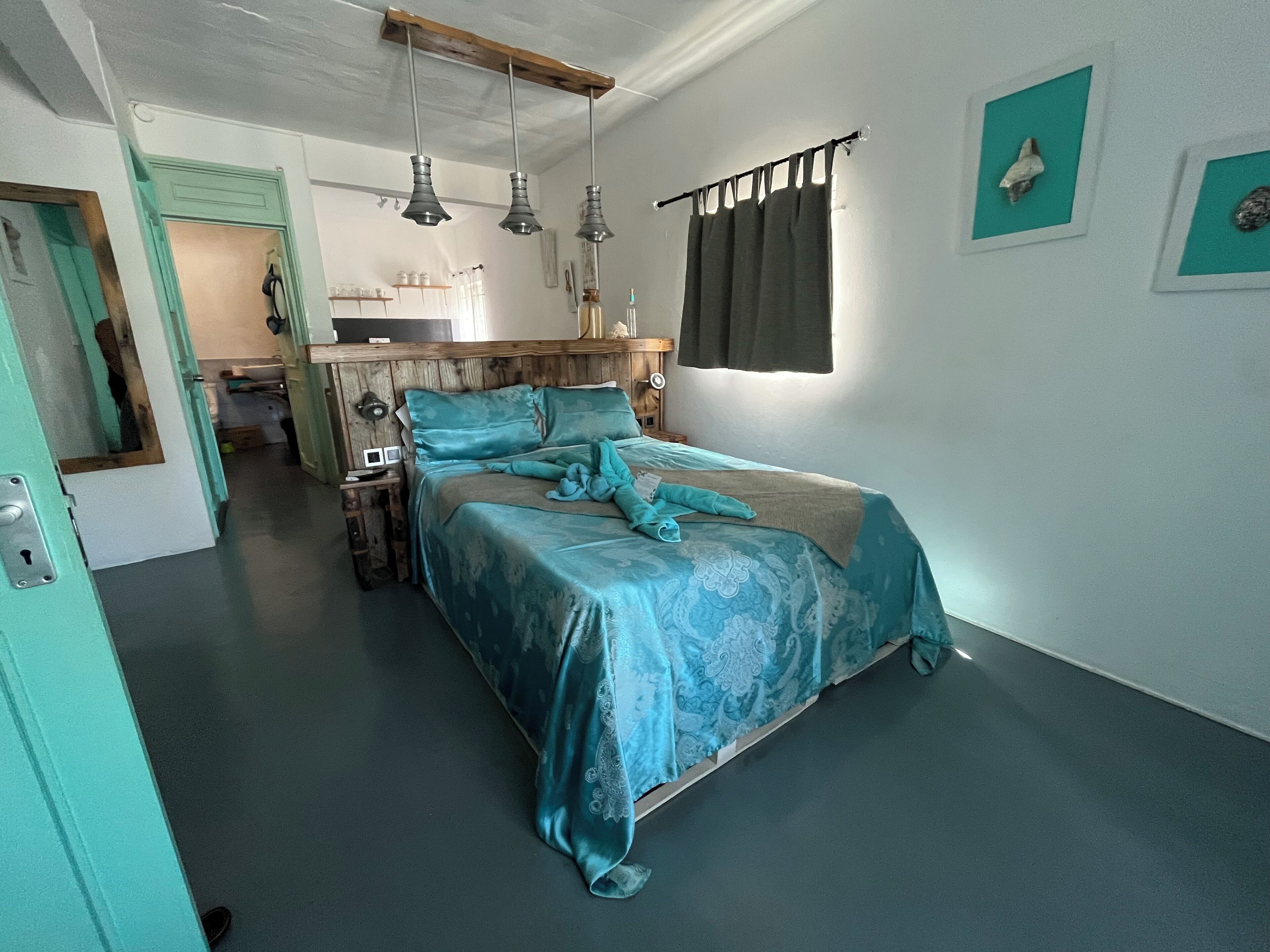 A bedroom with a bed, a wooden headboard, pendant lights, a window with curtains, and a small bathroom visible in the background.