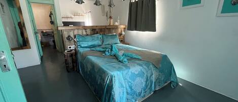 A bedroom with a bed, a wooden headboard, pendant lights, a window with curtains, and a small bathroom visible in the background.