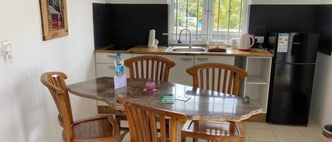 A compact kitchen with a dining table, four wooden chairs, a black refrigerator, and a window with a view of greenery.