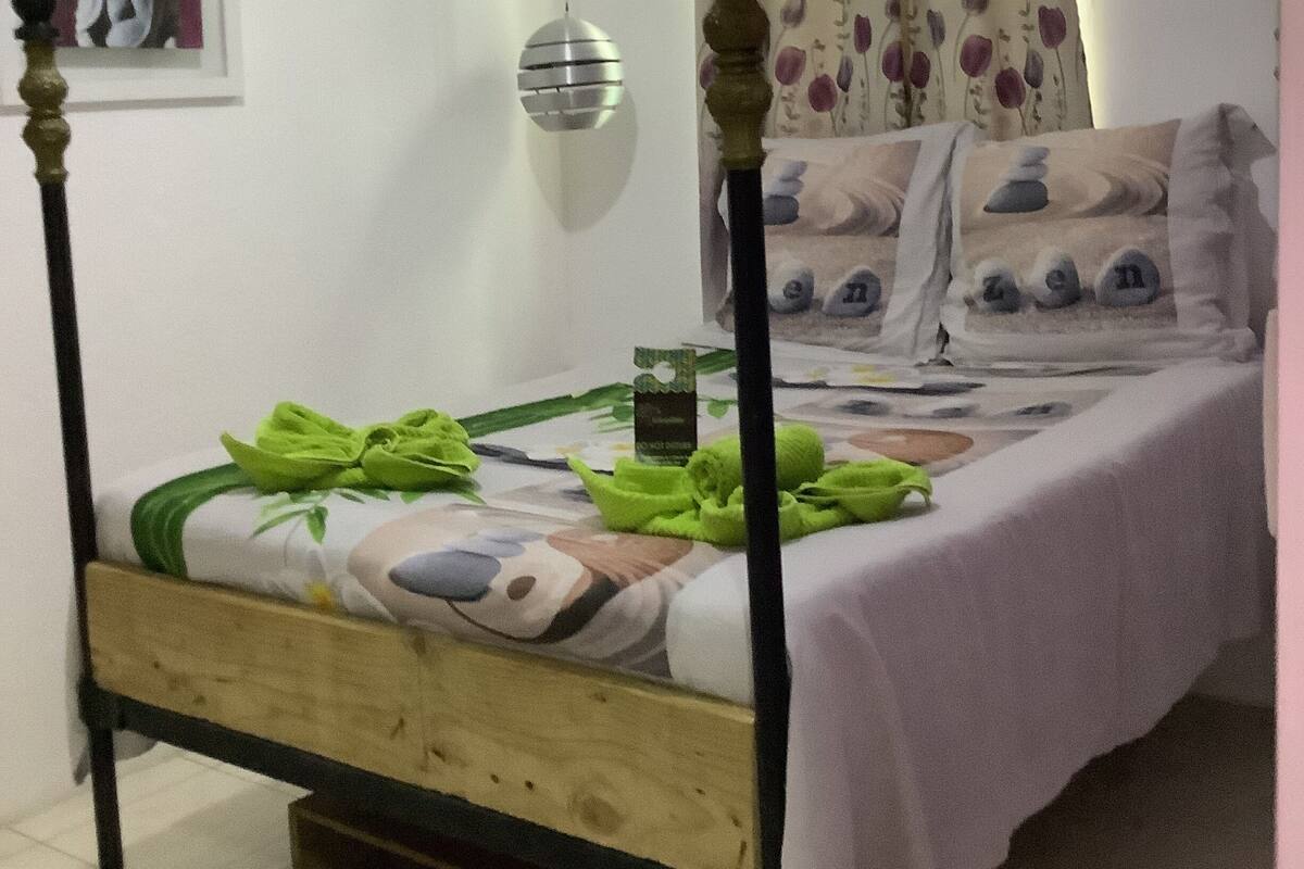 A four-poster bed with a wooden frame and a white bedspread, a small bedside table with a green cloth, and a wall-mounted lamp.