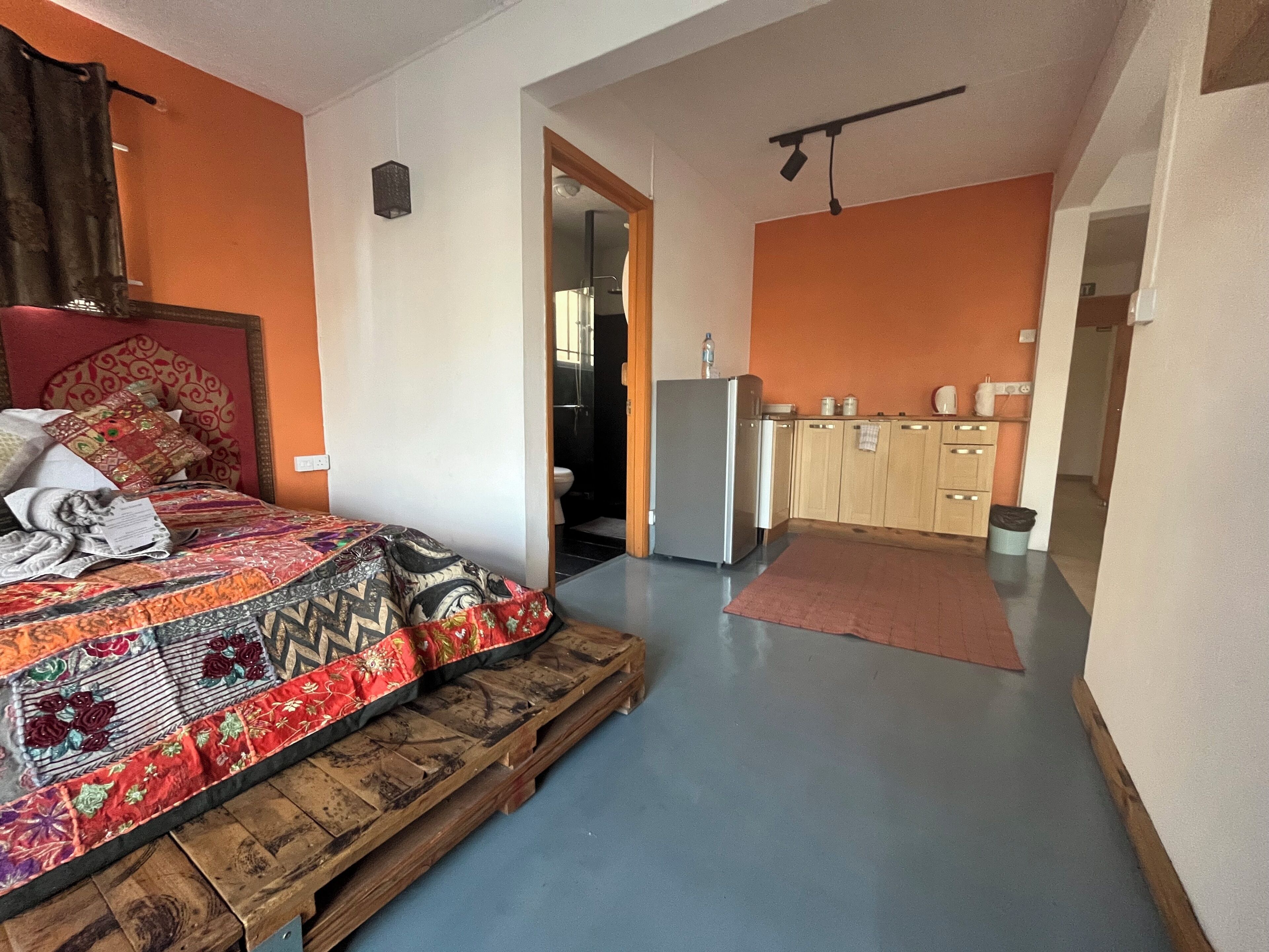 Een slaapkamer met een bed, een houten vloer, een keuken met kasten en een koelkast, en een badkamer zichtbaar door een geopende deur.