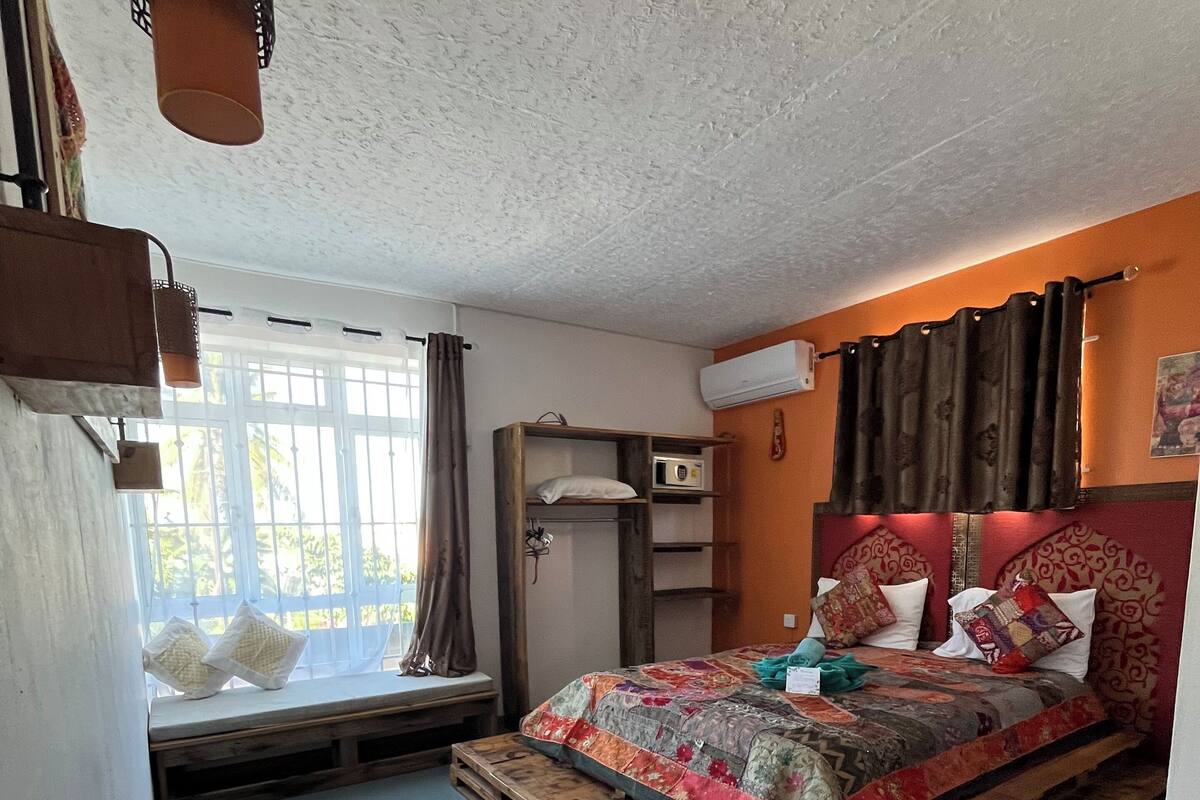 A bedroom with a bed on a wooden pallet, a window with curtains, a radiator, and a wall-mounted air conditioner.