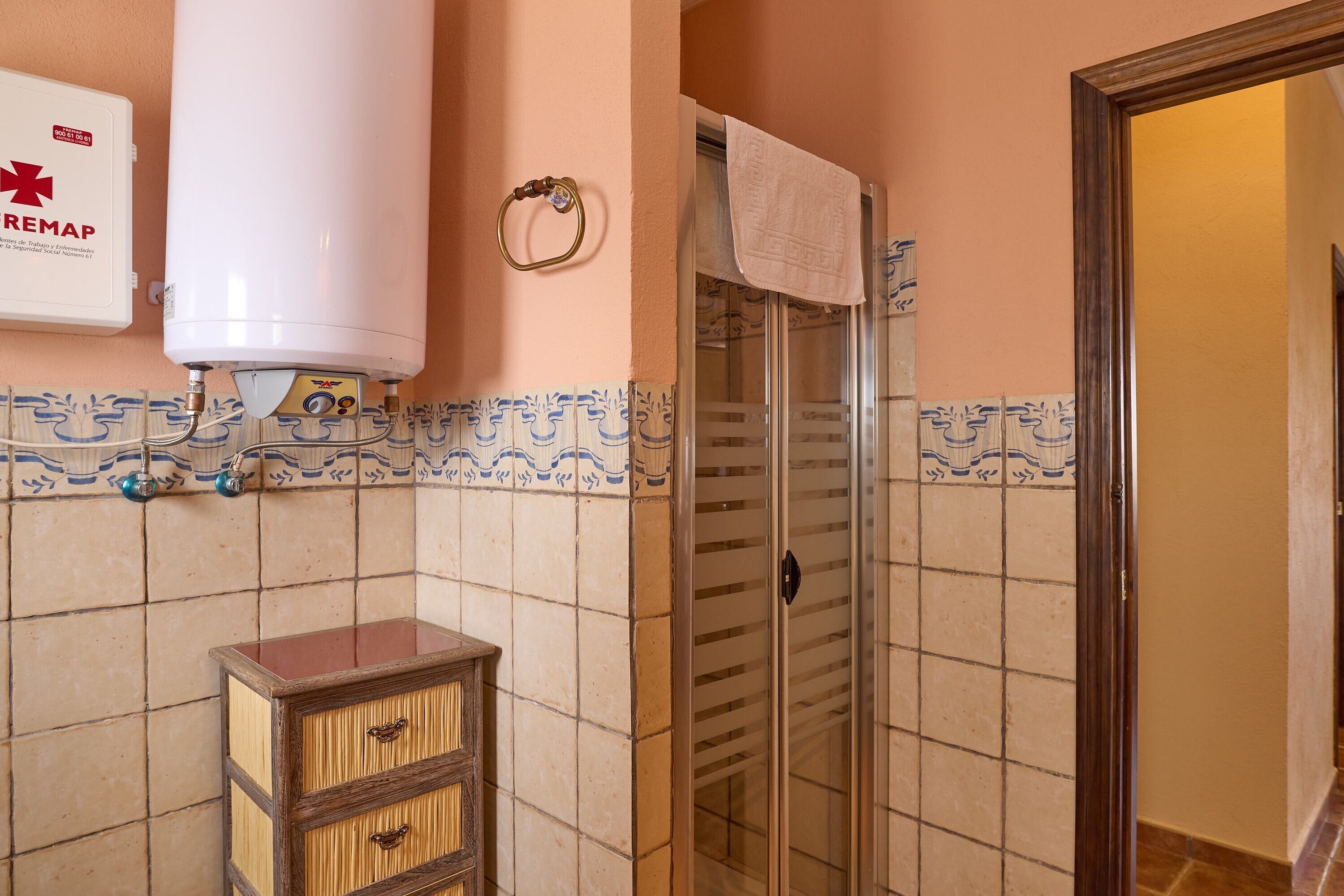 Casa de banho com esquentador branco, armário de madeira, cabine de duche em vidro e parede revestida de azulejos com padrão azul.
