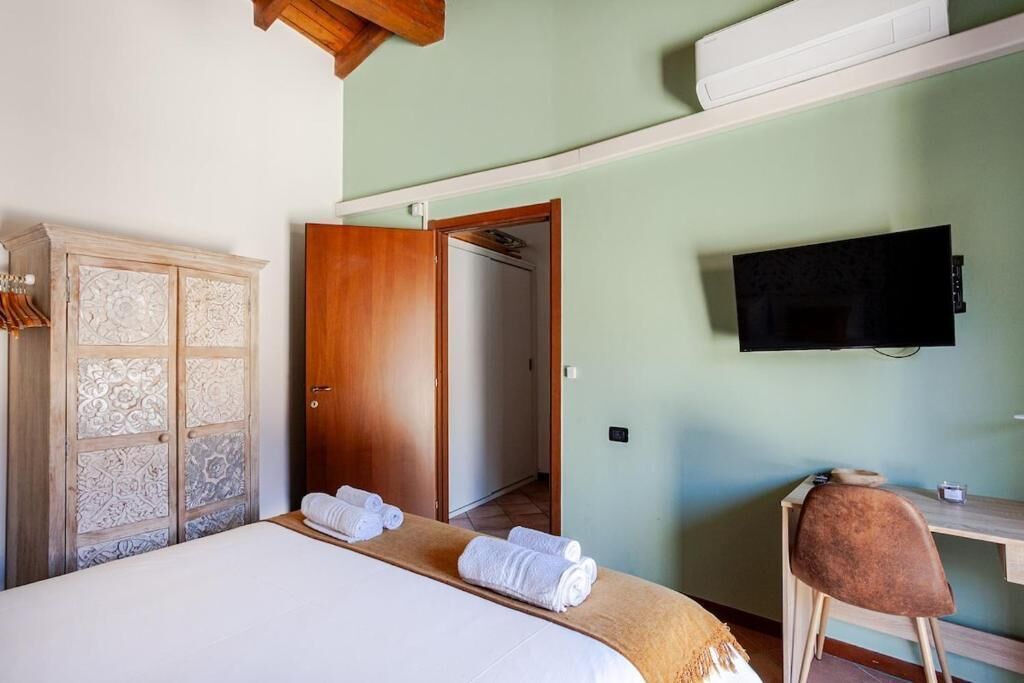 A bedroom with a bed, a wooden wardrobe, a desk, a chair, and a television on the wall.