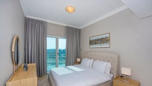 A hotel room with a large bed, a desk, a chair, a mirror, a painting, and a view of the city. - Large 1BR with Lake View in JLT  Close to Metro (Dubai)