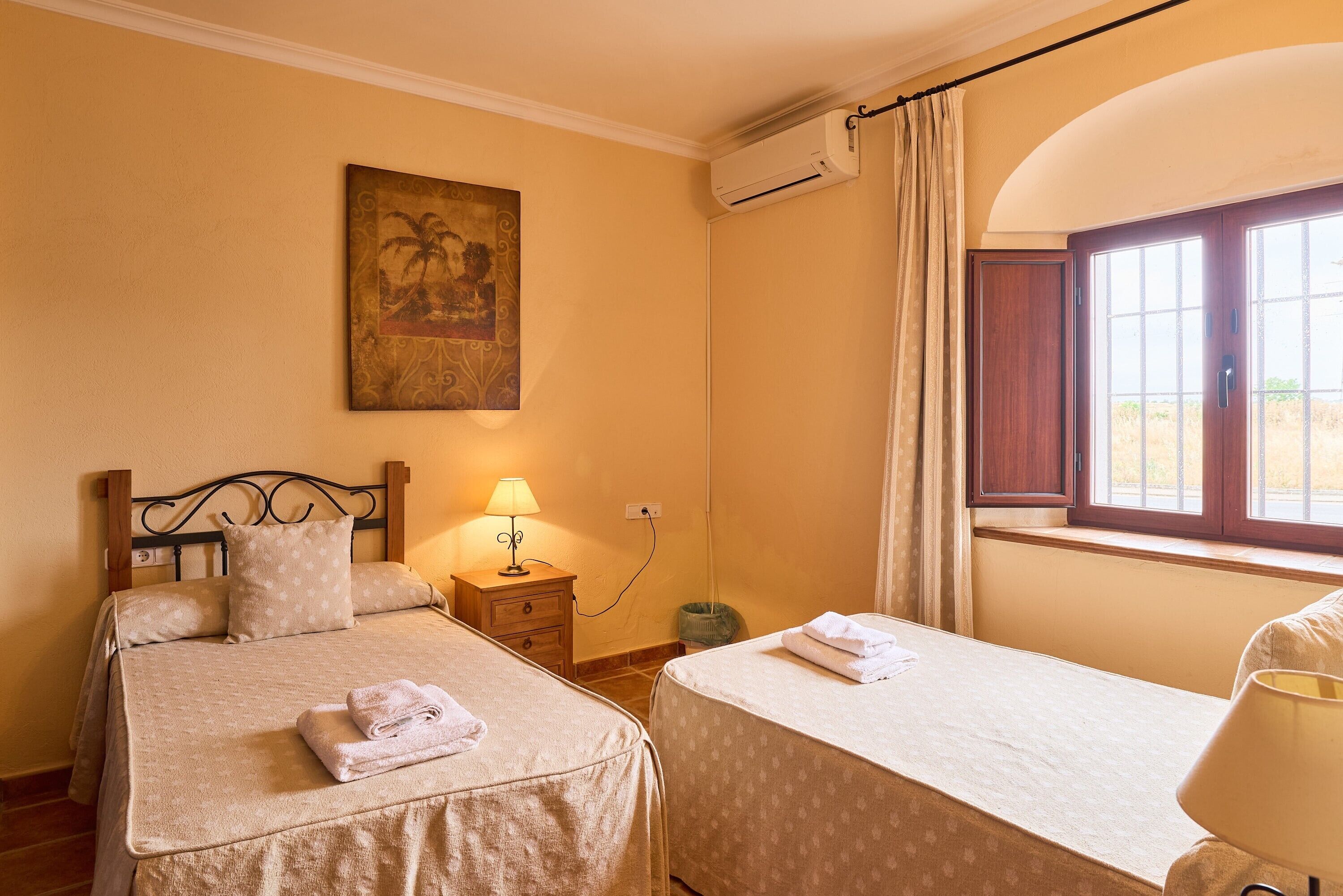Una habitación con dos camas, un cuadro en la pared, un equipo de aire acondicionado, una ventana con cortinas y una lámpara en una mesita de noche.