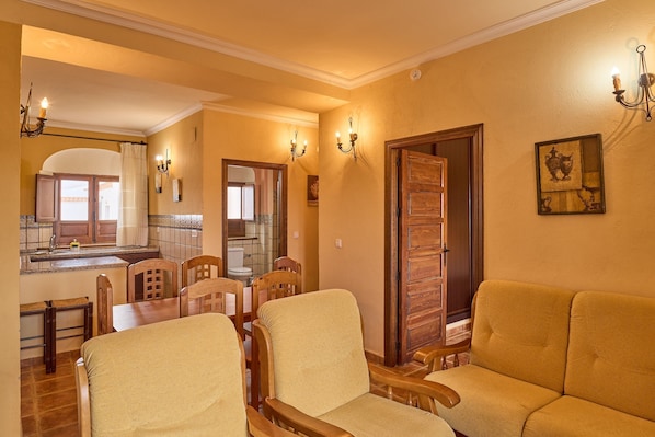 Un coin repas avec des chaises et une table en bois, un canapé, une cuisine avec une fenêtre et une porte donnant sur une autre pièce.