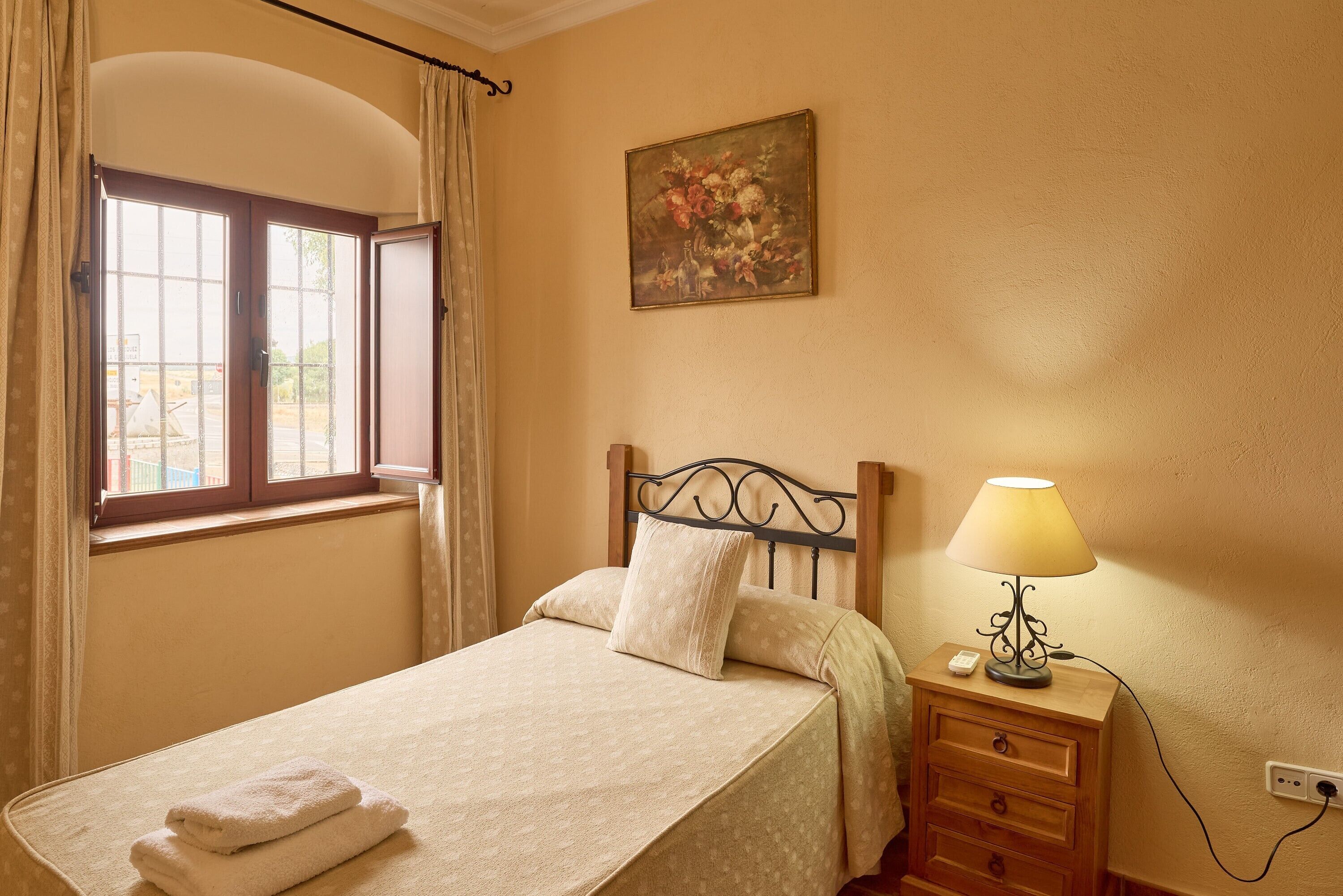 A bedroom with a bed, a nightstand, a lamp, a painting, and a window with curtains.