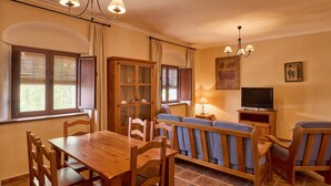 Un área de comedor con una mesa y sillas de madera, un sofá con cojines azules, un televisor sobre un mueble de madera y una araña.