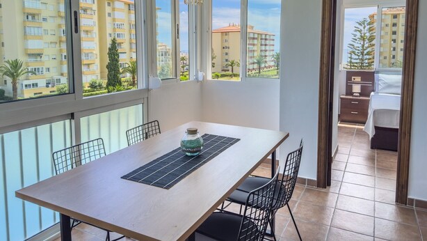 A dining area with a table, chairs, and a decorative bowl. There are large windows with a view of buildings and trees. - Apartment 'Sol and Sea A121' with Sea View, Wi-Fi and Air Conditioning (El Morche)