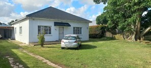 A white house with a dark roof, a car parked in the driveway, and a small tree in front. - Villiers Villa (Gqeberha)