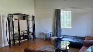A living room with a black leather sofa, a small round table, a white side table, a black metal shelf unit, and a window with curtains. - Villiers Villa (Gqeberha)