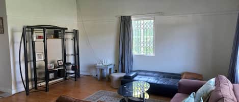A living room with a black leather sofa, a small round table, a white side table, a black metal shelf unit, and a window with curtains.