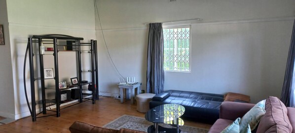 A living room with a black leather sofa, a small round table, a white side table, a black metal shelf unit, and a window with curtains. - Villiers Villa (Gqeberha)
