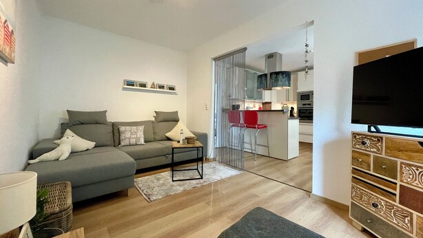 A modern living room with a grey sofa, a coffee table, and a TV. There is a kitchen area visible through a glass door. - 2-room apartment on the ground floor close to the beach (Büsum)